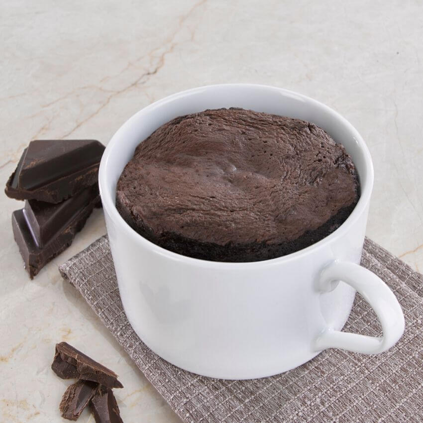 Delicious protein mug cake in a white cup, surrounded by chocolate pieces on a textured surface.